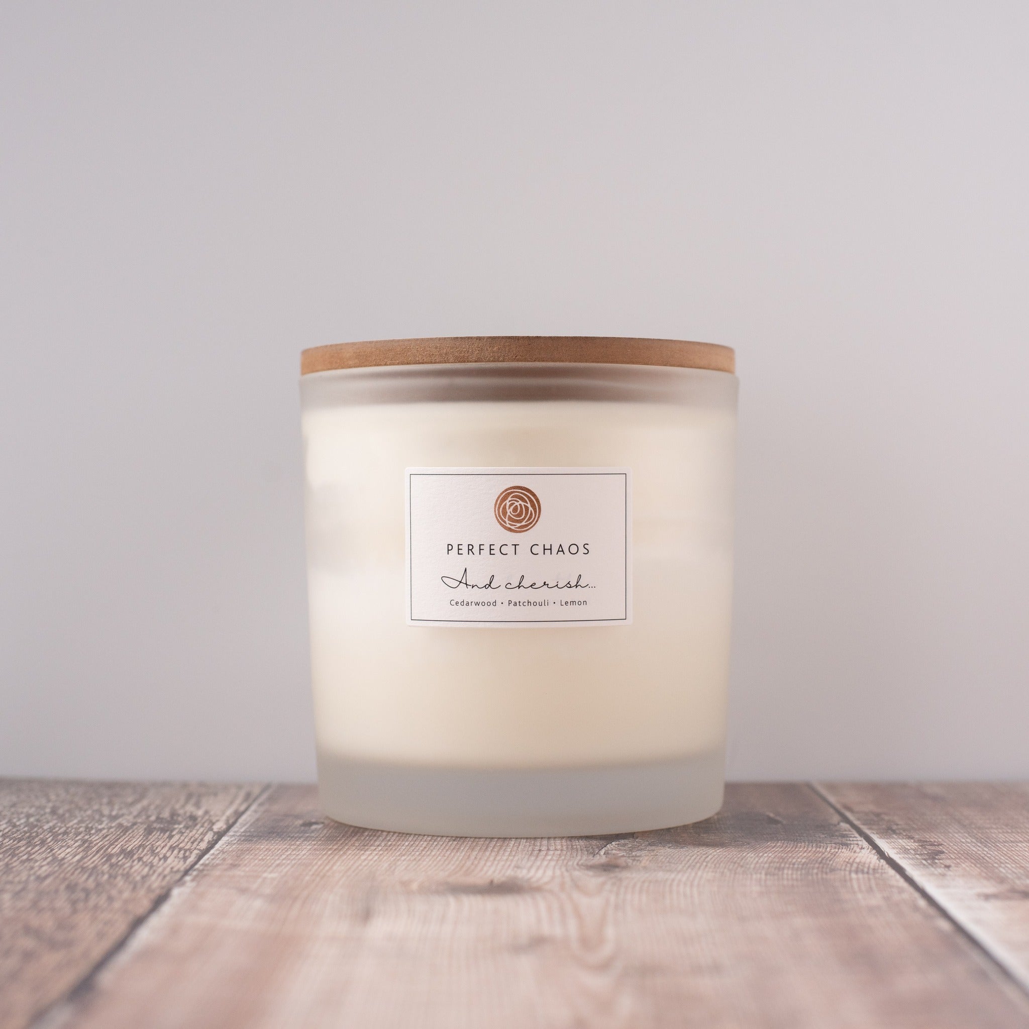 A frosted glass candle jar with wooden lid and perfect chaos label holding off-white wax scented with cedarwood patchouli lemon essential oils and long hours burn time