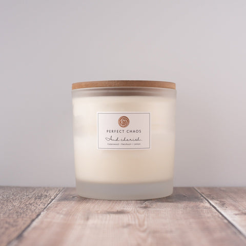 A frosted glass candle jar with wooden lid and perfect chaos label holding off-white wax scented with cedarwood patchouli lemon essential oils and long hours burn time