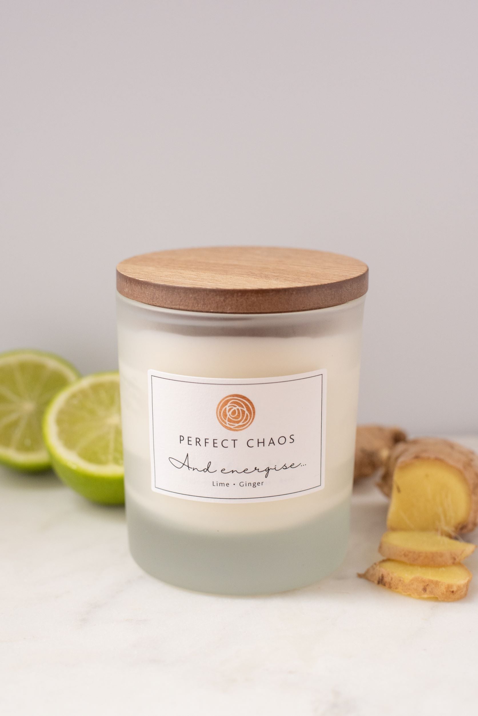 A frosted glass candle jar with wooden lid labeled perfect chaos and energise lime ginger with hours burn time