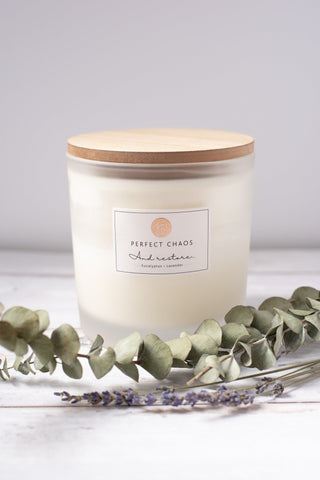 A creamy white frosted glass candle with wood lid labeled perfect chaos and rest featuring eucalyptus lavender essential oils and long hours burn time