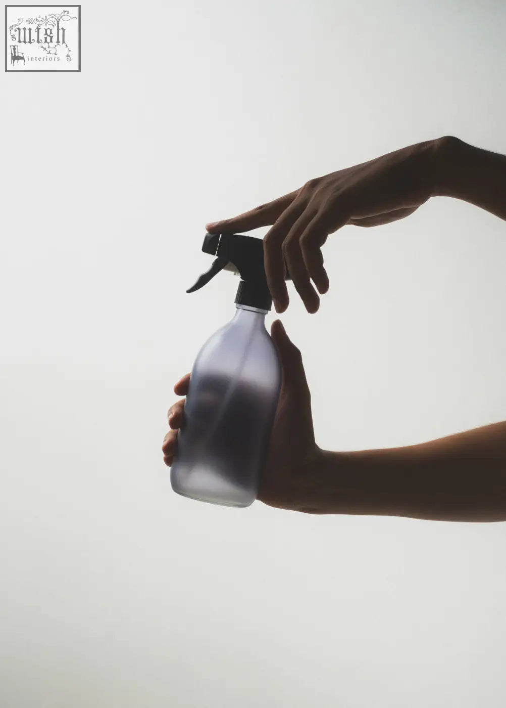 A frosted grey spray bottle with a black trigger pump held gently by two hands against a soft white background