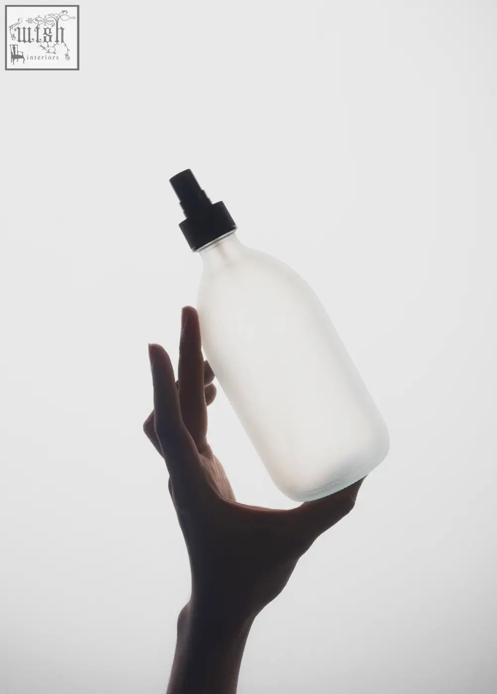 A frosted white atomiser bottle made of white glass held gently in a hand with a black spray nozzle against light background