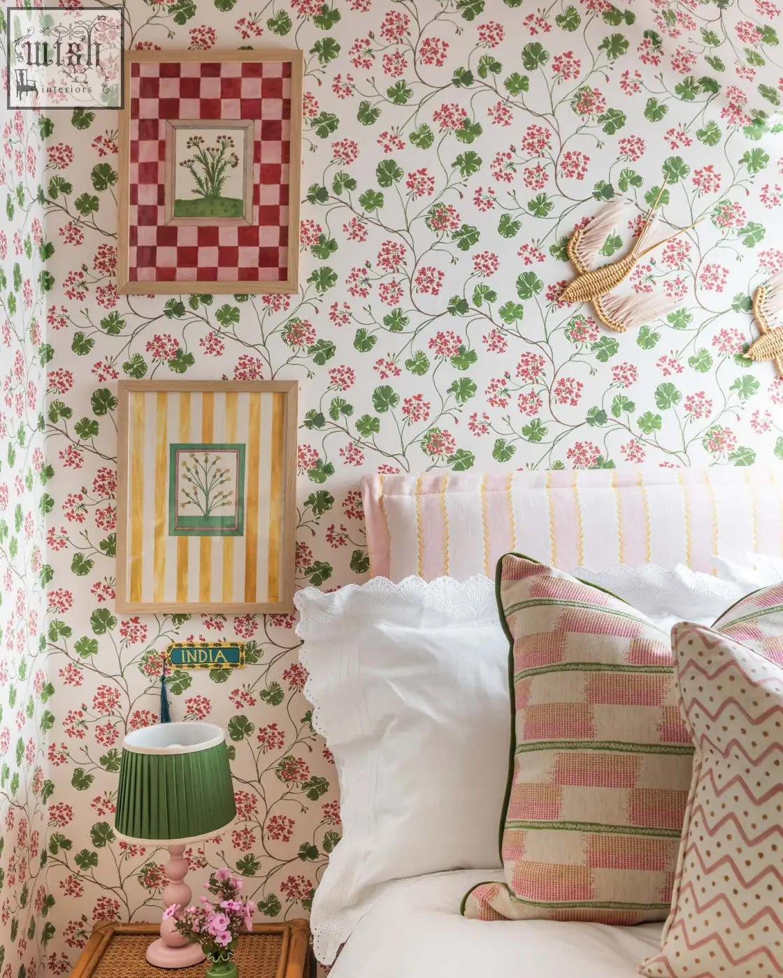 A green pleated lampshade with a pink base on a woven side table complements mediterranean charm with geraniums wallpaper