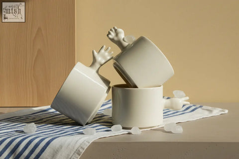 White ceramic gesture ceramic cups with sculpted hand handles stacked on striped cloth by 4am product design