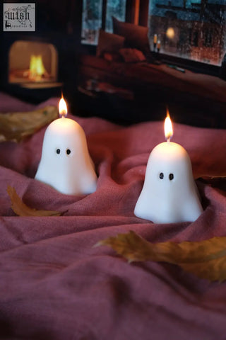 Two white ghost candles with black dot eyes lit on maroon fabric perfect for pumpkin spiced halloween decor