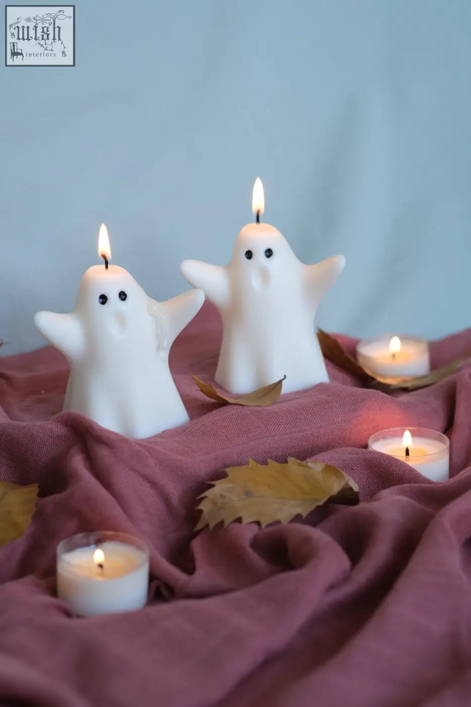 White ghost-shaped halloween candles with lit wicks on mauve fabric perfect for pumpkin spiced spooky vibes
