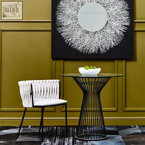 Black wireframe side table with glass top and white bowl of greenery next to gilden grey dining chair with woven back