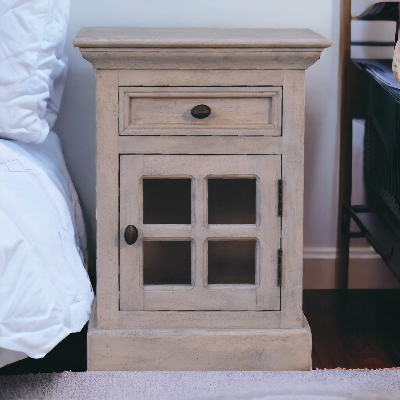 A rustic light gray wooden glazed stone bedside table with a drawer and glass-paneled door with dark metal hardware