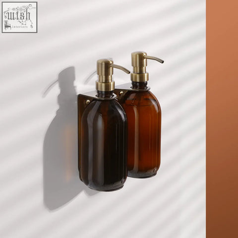 Gold double wall mounted soap dispenser with amber refillable glass bottles and brass pump nozzles on stainless steel bracket