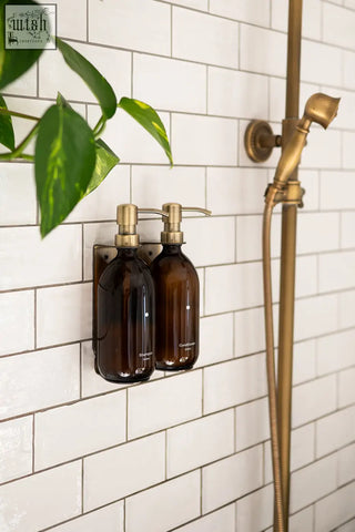 Two amber glass gold wall-mounted soap dispensers with brass pumps for shampoo and soap on a bathroom wall