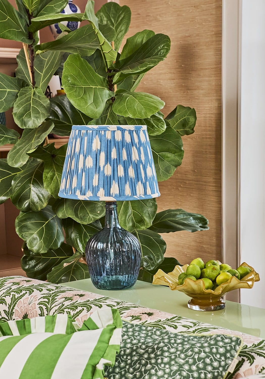 Blue glass table lamp with a pleated shade featuring white ikat-style patterns in a green cabana stripe ruffled cushion cover
