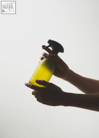 A translucent yellow glass spray bottle with a black trigger nozzle held in two hands against a bright background