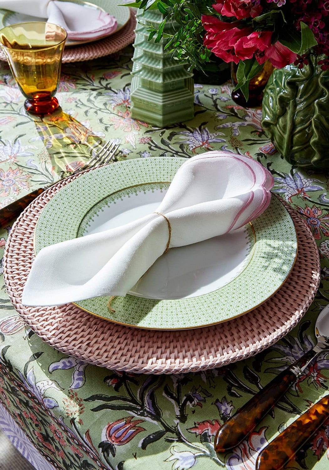 A white napkin with a pink edge tied with gold on a green with envy floral print tablecloth