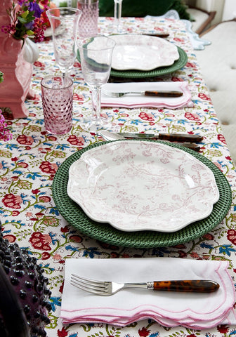 A delicate white ceramic plate with pink floral pattern on a green woven harbour island round rattan charger set of 4