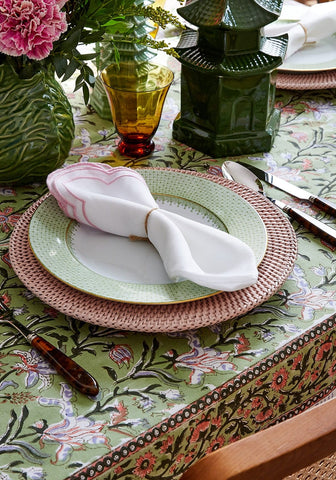 A white napkin with a pink border tied with a gold tie on a harbour island round rattan charger placemat pink set of 4