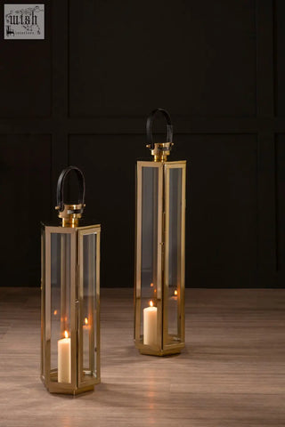 Two elegant medium gold finish lanterns with clear glass panels and black leather handles each holding a lit candle
