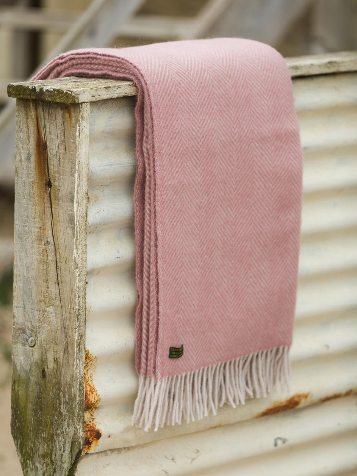 A dusky pink pearl herringbone pure new wool throw with fringed edges draped over rustic wood and metal surfaces