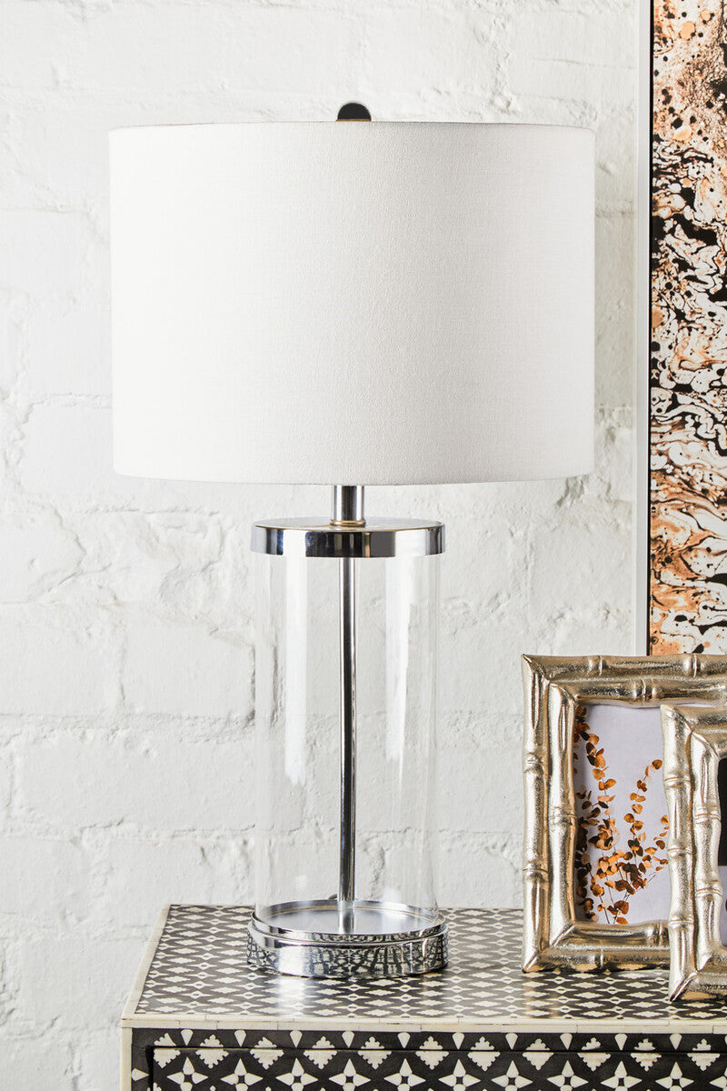 Clear glass cylinder table lamp with white fabric shade and chrome accents next to a silver finish bamboo effect photo frame
