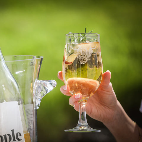 A clear glass highland cow craft beer goblet with a delicate stem holding a golden craft beer with ice and rosemary