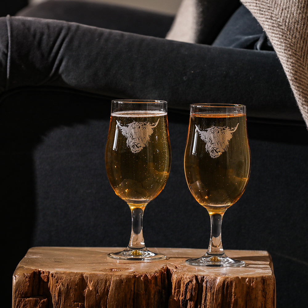 Two clear glass craft beer glasses with etched white highland cow design filled with golden liquid