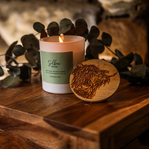 A lit pale pink soy wax candle in a white glass jar with wooden lid engraved with a highland cow and fresh fennel scent