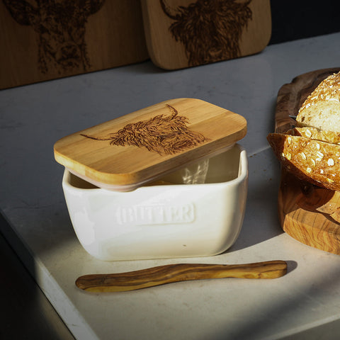 White ceramic butter dish with wooden lid engraved with highland cow design and olive wood spreader beside it