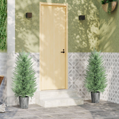 Two tall green artificial cypress trees with feathery foliage in gray metal pots flanking a light wood door