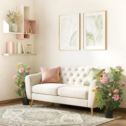 White tufted velvet sofa with gold tone legs and pink green pillows next to a pink artificial rose tree set for indoor decor