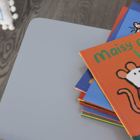 A stack of colorful maisy kids board books on an orange cover beside a grey homcom kids table and chair set for toddlers
