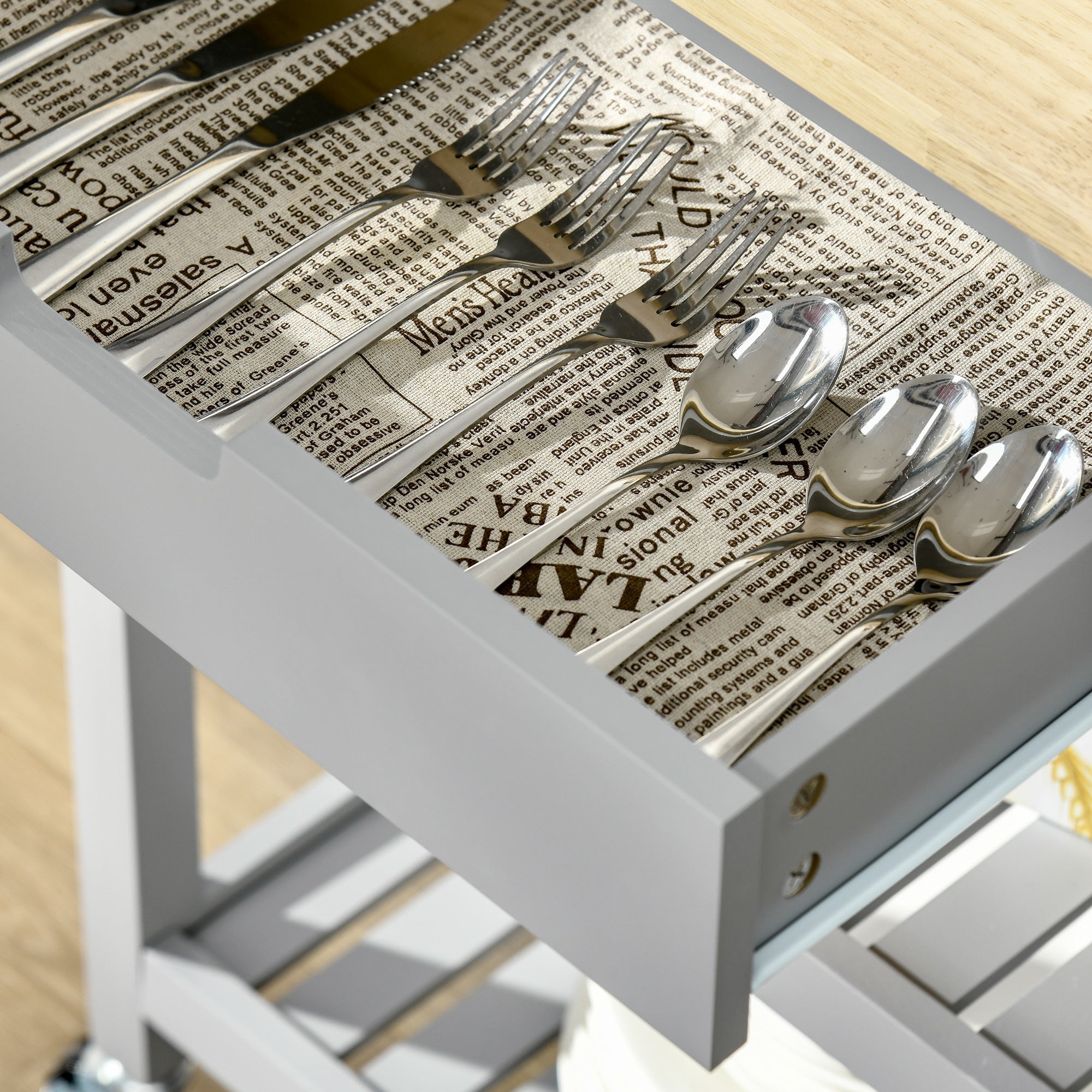 Shiny stainless steel cutlery set neatly arranged on newspaper-lined drawer of grey homcom kitchen trolley with towel rack