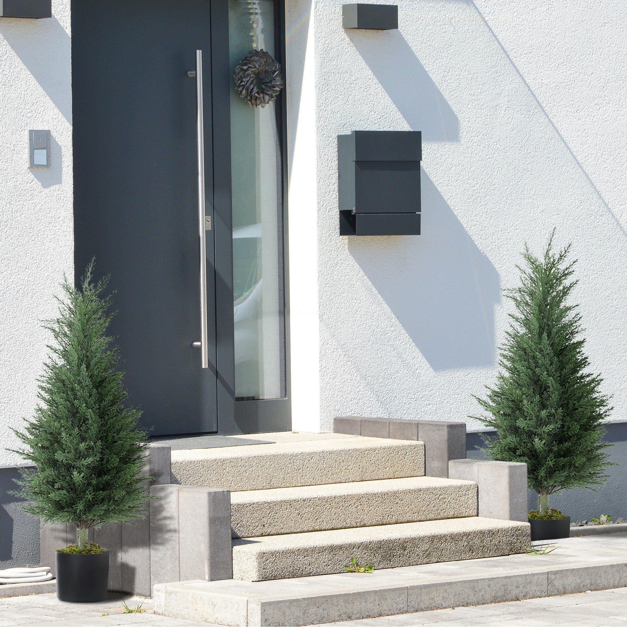 Black modern mailbox mounted on white stucco wall next to homcom artificial cypress tree in nursery pot for green thumb decor