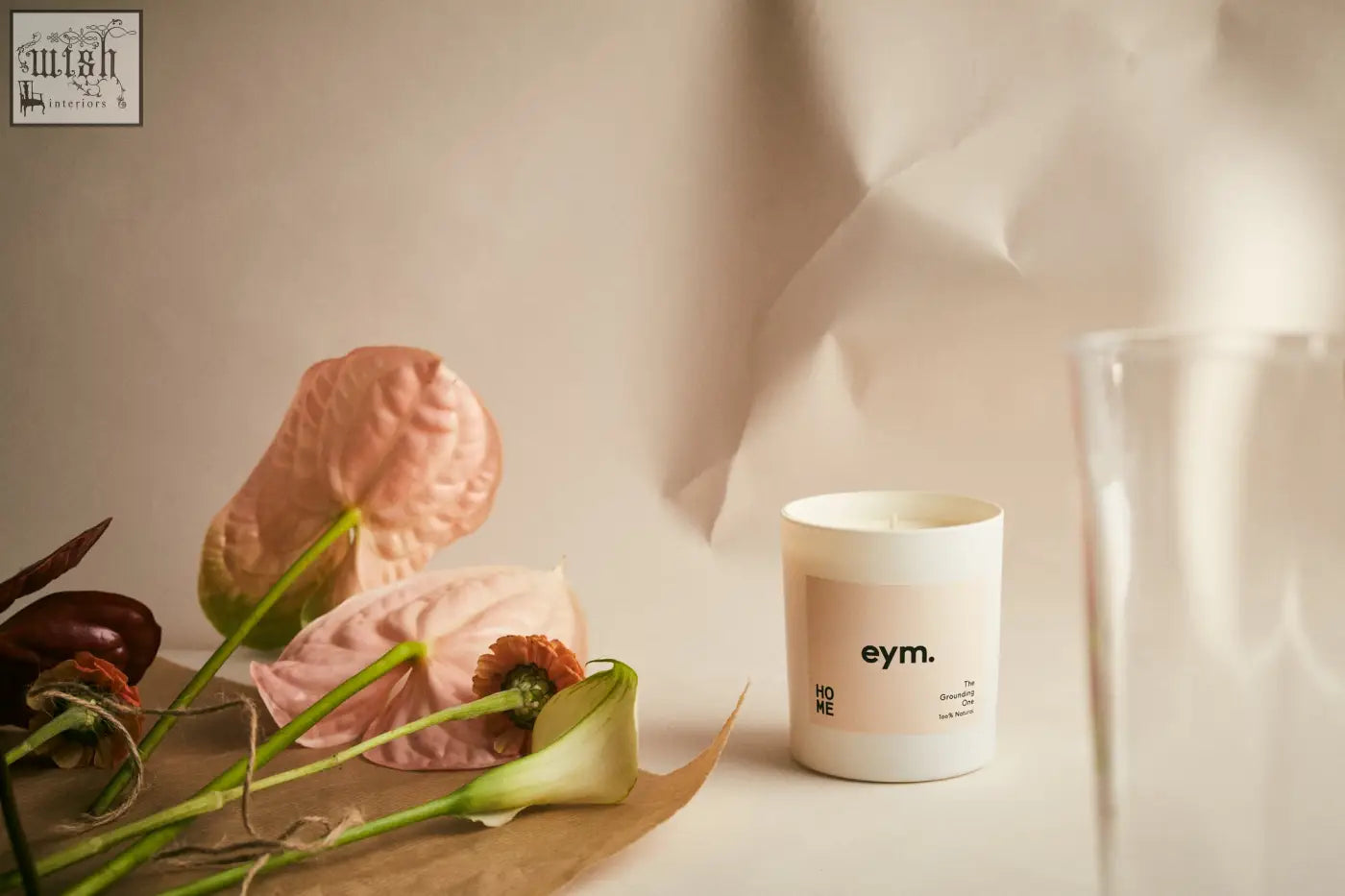 A minimalist white standard candle jar with a matte finish and beige eym label next to soft pink flowers on brown paper