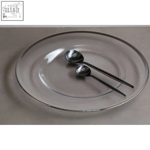 Clear glass charger plate with silver rim holding two black spoons for elegant table setting
