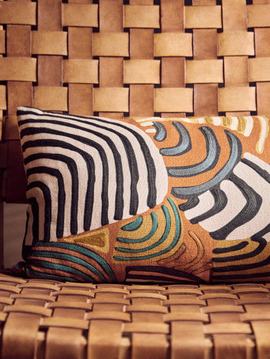 A rectangular cushion with bold black white stripes and orange teal swirl patterns on the inca armchair with leather straps and american oak frame