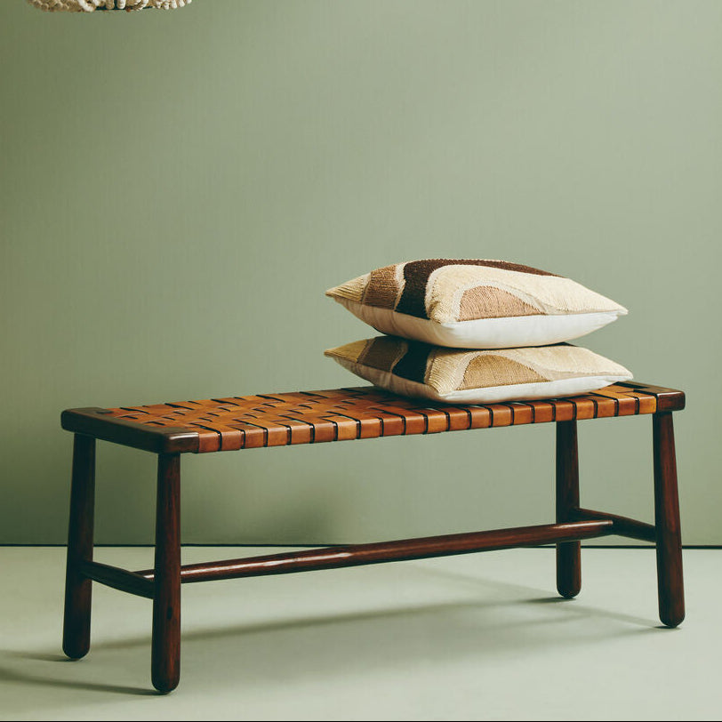 A rustic strapped bench with tan leather woven seat and dark mango wood legs topped with beige patchwork pillows