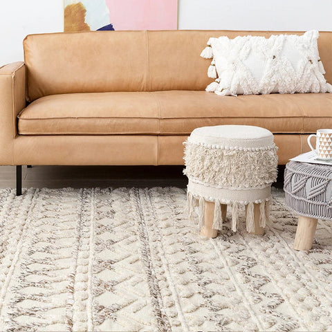 Cream-colored leather sofa with a textured white pillow and matching woven ottoman next to Initoma rug