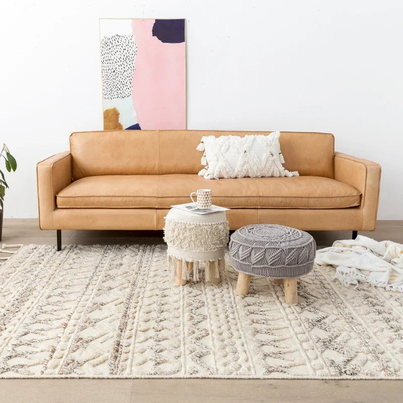 Tan leather sofa with black legs and a white textured pillow shown with initoma rug in the background