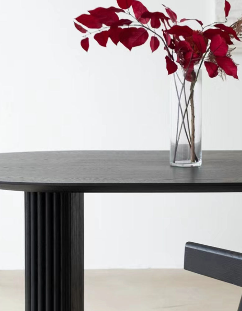 A sleek black ivar oval dining table with a fluted column base and a clear glass vase holding red leaves