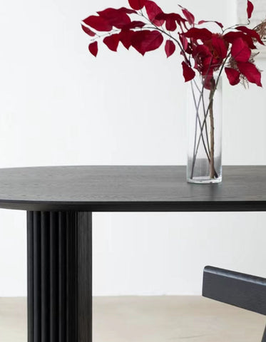 A sleek black ivar oval dining table with a fluted column base and a clear glass vase holding red leaves
