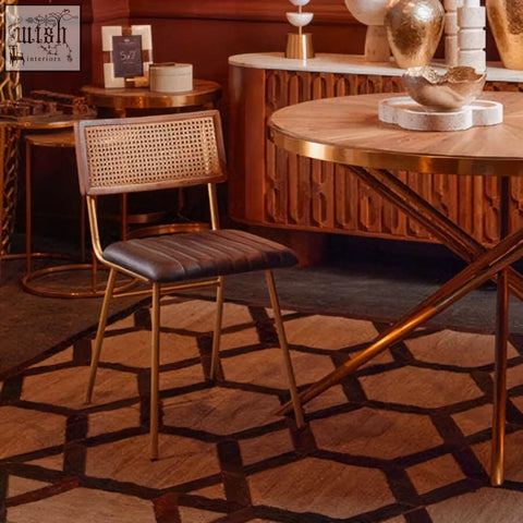 Gold-finished metal chair with woven cane backrest and dark brown leather seat next to jana round fir wood dining table