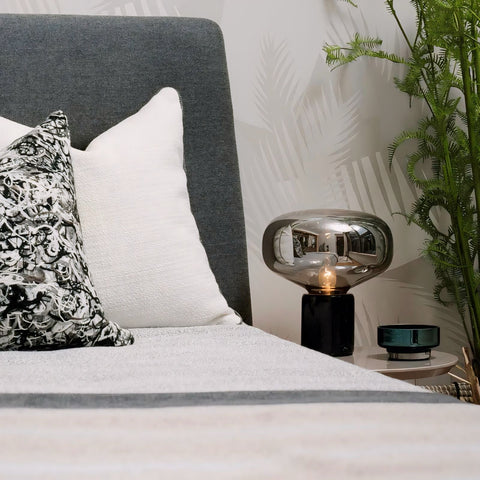 A sleek silver glass johano light with a reflective surface and black base sits on the bedside table