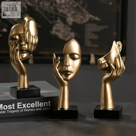 Three golden metallic sculpted face resting ornaments with hand gestures on black bases kanso ornament