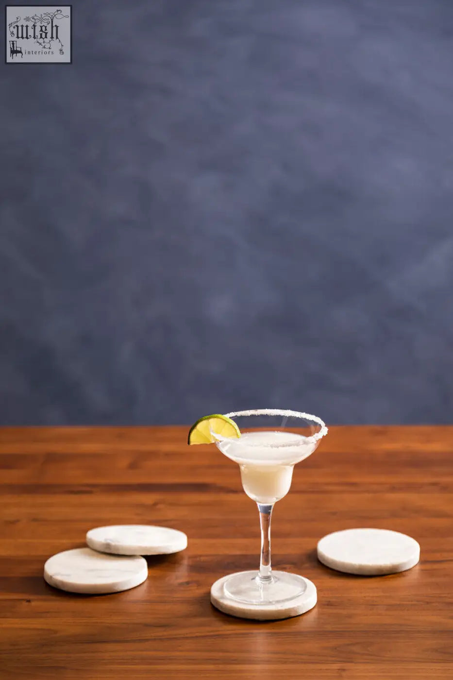 A margarita served on a clear glass with a lime wedge on a set of 4 white marble round coasters in kindia style