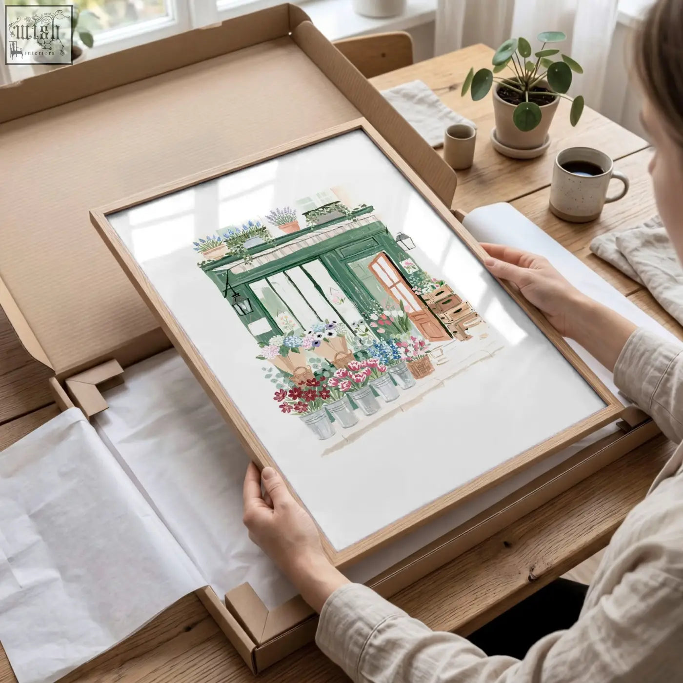 A framed watercolor kitchen print of a green country cottage flower shop with colorful blooms in a light wood frame