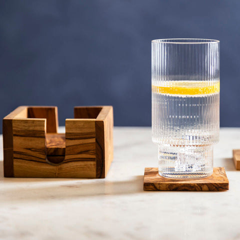 Clear ribbed glass tumbler with lemon slice resting on olive wood square coasters from kora set of 4