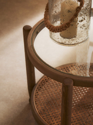 A rustic two-tier side table with glass top and woven cane shelves framed by dark wood legs featuring natural rope details