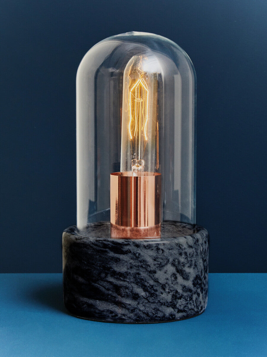 A lamonte bell lamp with a warm glowing edison bulb under a clear glass dome on a polished black marble base
