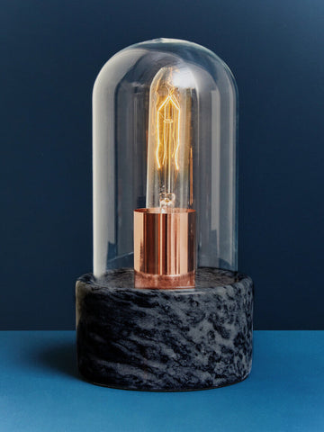 A lamonte bell lamp with a warm glowing edison bulb under a clear glass dome on a polished black marble base