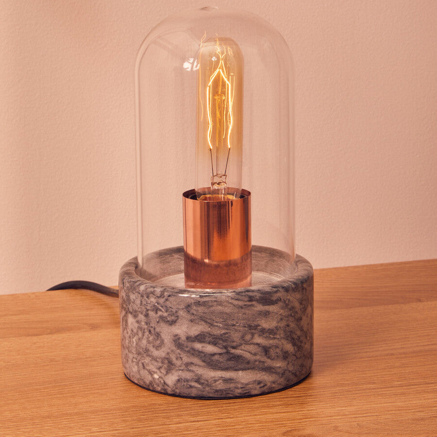 A warm-toned lamonte bell lamp with a clear glass dome and black marble base featuring a copper holder and filament bulb