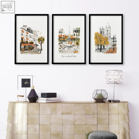 Three black-framed large artwork prints featuring autumn paris watercolour scenes hang above a gold textured console in a living room